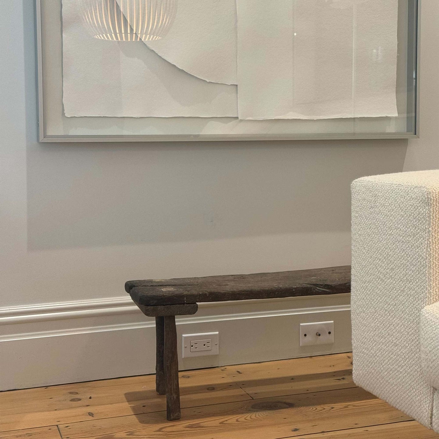 Partial side view of antique small table made of dark brown wood in a room with white background and hardwood floors. An all white framed work of art with the reflection of a round, white light fixture rests on the background wall behind the table. A white, woven textured couch sits partially in front of the table.